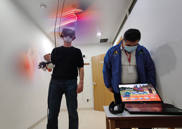A man stands wearing a VR headset and holding a controller, next to a man looking at a game screen on a laptop A man stands wearing a VR headset and holding a controller, next to a man looking at a game screen on a laptop