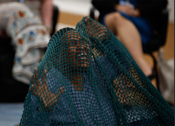 An African woman kneels with hands in the air as if in pain, covered with a large blue fishing net, surrounded by an audience An African woman kneels with hands in the air as if in pain, covered with a large blue fishing net, surrounded by an audience