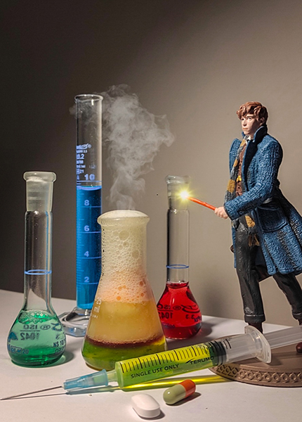 A figurine of a man with a wand stands to the right of various beakers filled with colourful liquids with smoked emerging, and a syringe and tablets in the foreground. A figurine of a man with a wand stands to the right of various beakers filled with colourful liquids with smoked emerging, and a syringe and tablets in the foreground.
