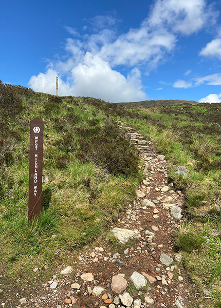 A rocky pathway leading up a grassy hill, with a sign saying the West Highland Way on the left A rocky pathway leading up a grassy hill, with a sign saying the West Highland Way on the left