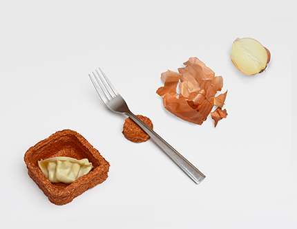A slice of onion next to crumpled onion skins, a fork next to a little rough-textured container holding a gyoza A slice of onion next to crumpled onion skins, a fork next to a little rough-textured container holding a gyoza