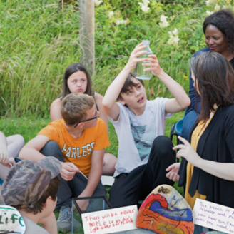 Group of young people sitting on grass with teachers while one holds up a bottle looking into it Group of young people sitting on grass with teachers while one holds up a bottle looking into it