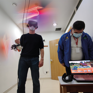 A man stands wearing a VR headset and holding a controller, next to a man looking at a game screen on a laptop A man stands wearing a VR headset and holding a controller, next to a man looking at a game screen on a laptop