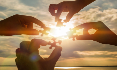 four hands putting together a jigsaw puzzle against a sunset sky